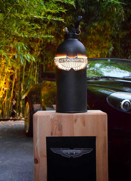 Lampe extincteur aston martin sur un socle en bois centré devant une aston martin JLR Concept